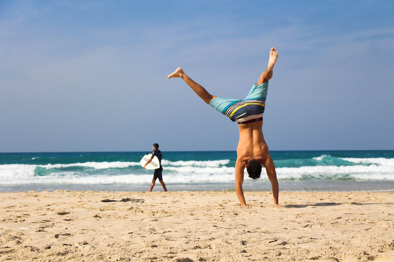 Man doing a handstand on a sunny beach with surfer in background, evoking a sense of freedom and joy.